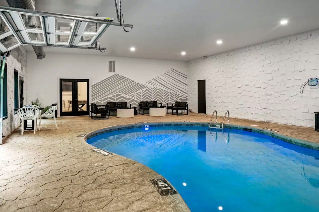 An indoor swimming pool with a geometric wall design, seating area with chairs and tables, and a visible door.