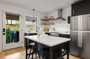 A modern kitchen with a central island, stainless steel appliances, and a view of a garden through a sliding door.