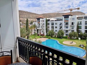 Un balcón de hotel con vista a una piscina, un jardín ajardinado y edificios de varios pisos al fondo.