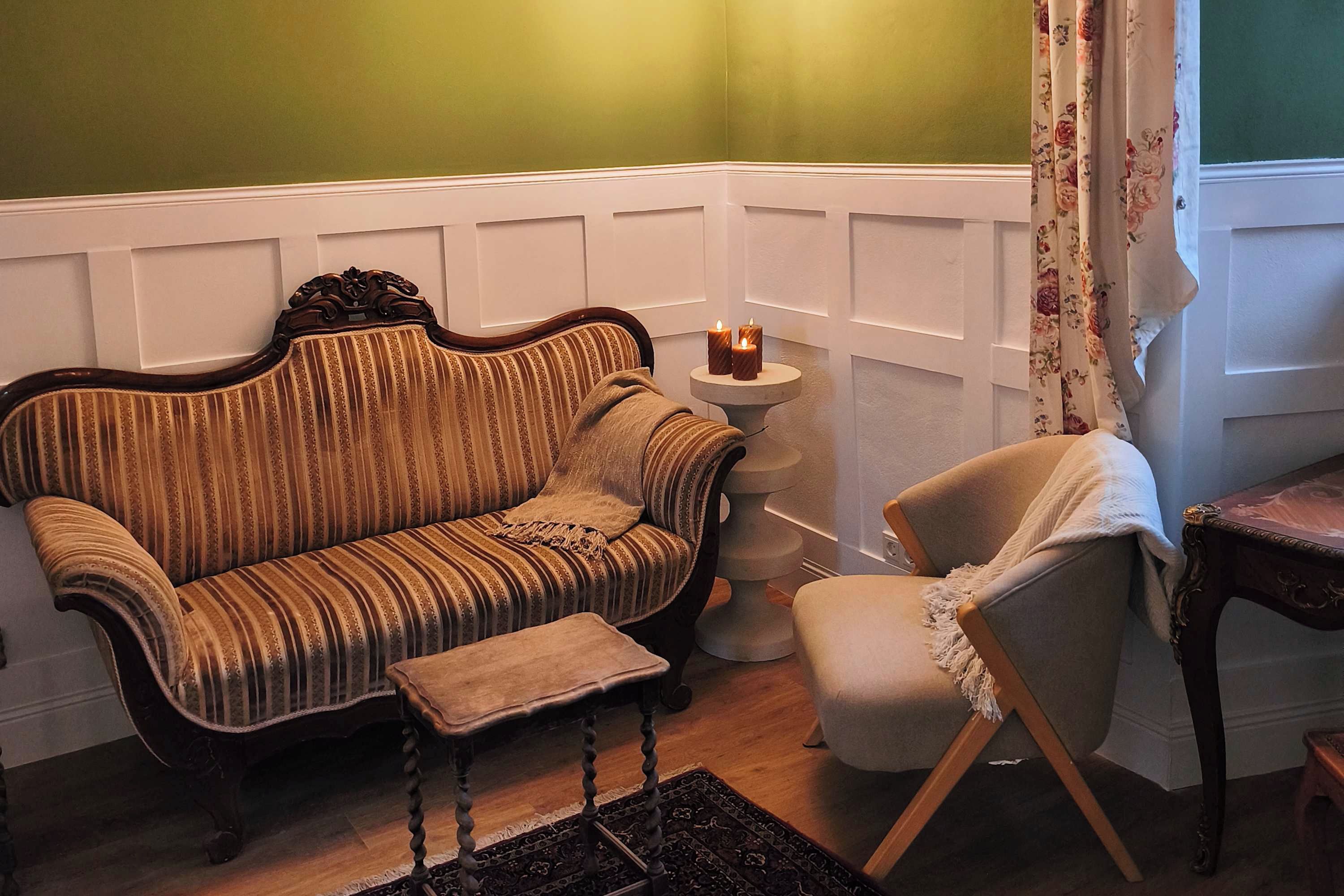Ein Eckzimmer mit einem Vintage-Sofa mit Streifen, einem kleinen runden Tisch mit Kerzen und einem schlichten Sessel.