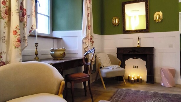 Una sala de estar acogedora con chimenea, un sillón cómodo, una mesita con una vela y una ventana con cortinas florales.