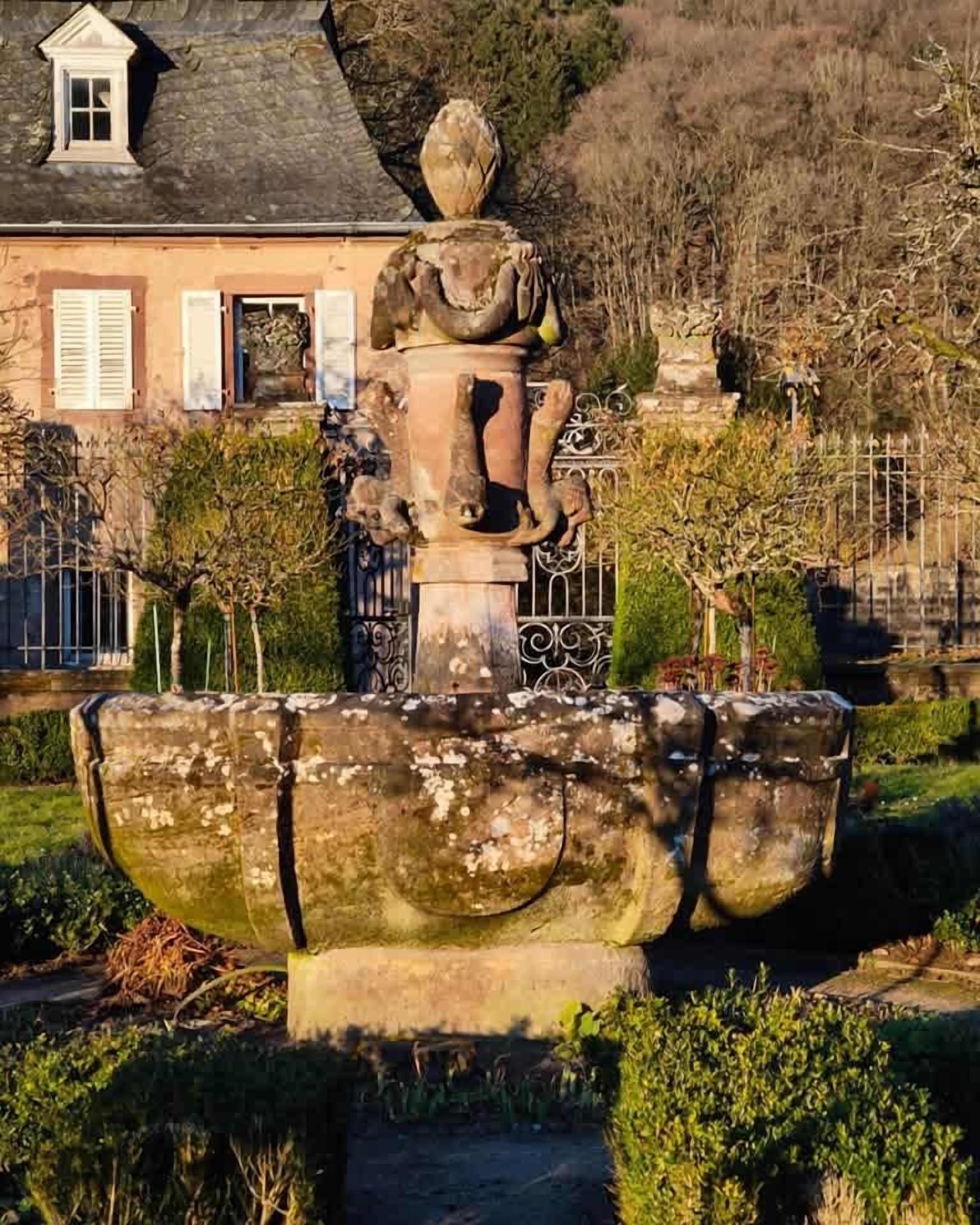 Una fuente de diseño clásico, rodeada de vegetación cuidada y un edificio con techo a dos aguas al fondo.