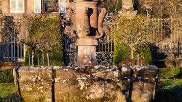 Una fuente de diseño clásico, rodeada de vegetación cuidada y un edificio con techo a dos aguas al fondo.