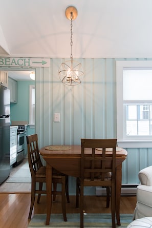 A dining area with a wooden table and chairs, a hanging light fixture, and a kitchen in the background. - Classic Maine Beach Cottage! (Scarborough)