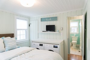 A bedroom with a bed, a washing machine, a television, and a bathroom visible through an open door. - Classic Maine Beach Cottage! (Scarborough)