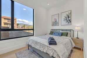 Een slaapkamer met een groot raam, een bed met beddengoed met een patroon, twee kunstwerken aan de muur, een bedlamp en een klein nachtkastje.
