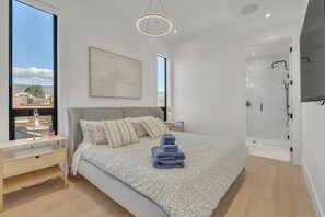 A modern bedroom with a large bed, a bedside table, a nightstand, a wall-mounted artwork, a shower area, and a view of the outside. - Lakeshore Luxury 3BR+Flex Space | Beachfront Condo (Penticton)