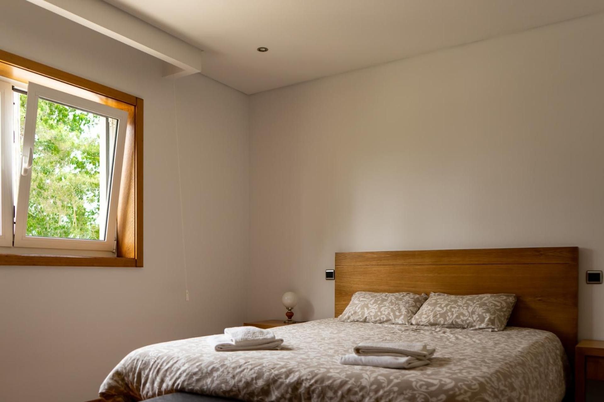 A bedroom with a wooden headboard, a bed with patterned bedding, a window with curtains, and a bedside table with a lamp.