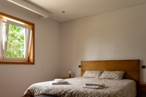 A bedroom with a wooden headboard, a bed with patterned bedding, a window with curtains, and a bedside table with a lamp.