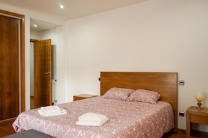 A hotel room with a wooden headboard, a bed with a patterned bedspread, two bedside tables, and a nightstand with a lamp.