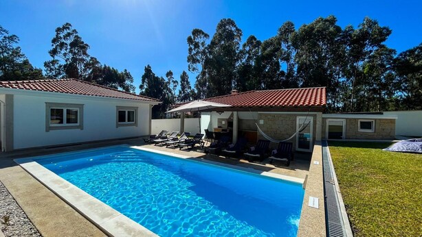 Una piscina con sillones de descanso y sombrillas, rodeada por una casa con techo de tejas y un patio con césped.