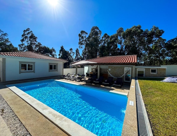 A swimming pool with lounge chairs and umbrellas, surrounded by a house with a tiled roof and a grassy yard.