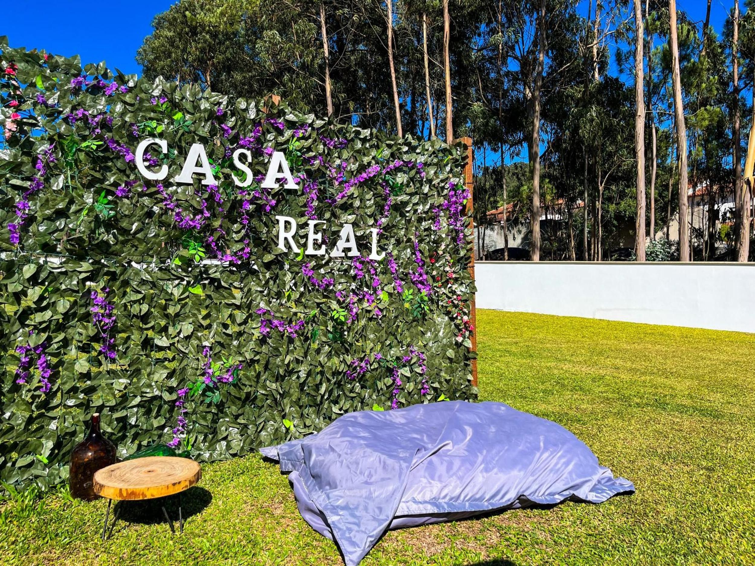 Un letrero con el texto 'CASA REAL' sobre un fondo de follaje verde y flores púrpuras.