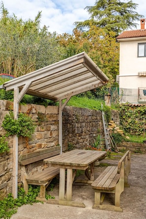 Eine hölzerne Pergola mit Bänken, vor einer Steinmauer und von Grünpflanzen umgeben.