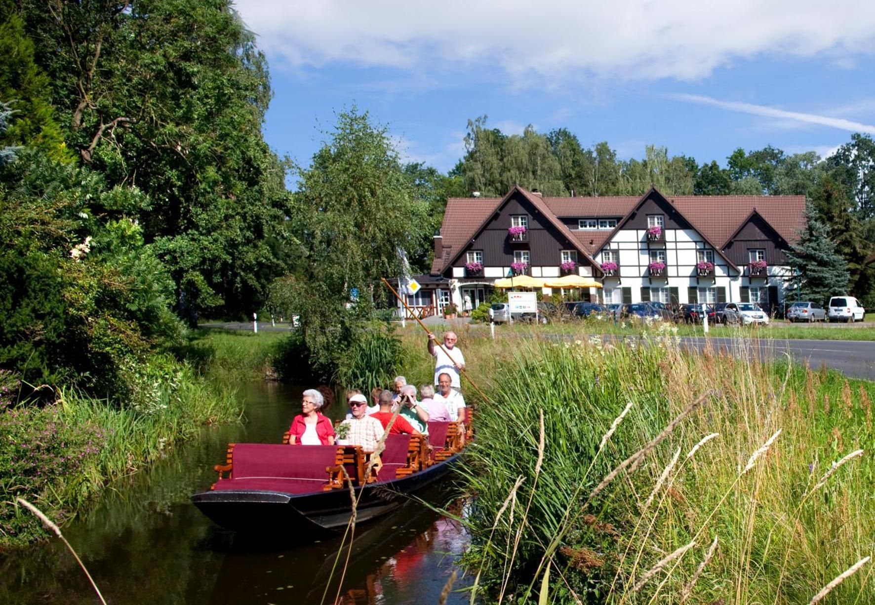 Ein schmaler Kanalboot mit Passagieren auf einem ruhigen Fluss, umgeben von üppiger Vegetation und einem Gebäude mit traditioneller Architektur im Hintergrund.