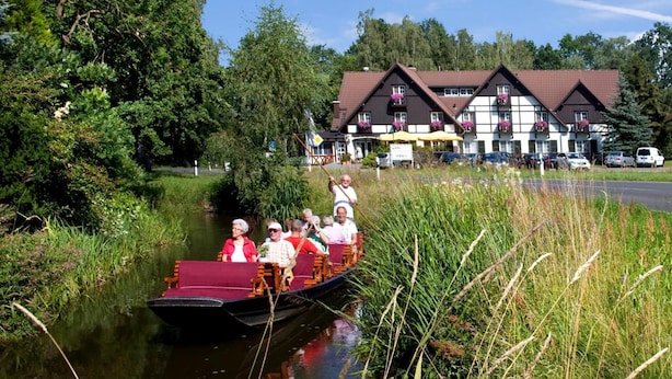 A narrowboat with passengers on a calm river, surrounded by lush greenery and a building with a traditional design in the background. - Hotel Am Spreebogen (Burg (Spreewald))