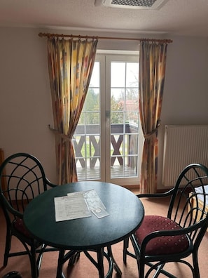 A room with a round table and two chairs, a window with curtains, and a view of a deck outside. - Hotel Am Spreebogen (Burg (Spreewald))