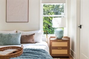 A bedroom with a bed, pillows, a bedside lamp, a wicker basket, and a view of greenery outside. - Beltline Bungalow (Atlanta)