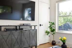 A modern living room with a flat-screen TV mounted on the wall, a dark wooden cabinet with books, a potted plant, and a window with a view of trees. - Beltline Bungalow (Atlanta)