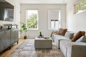 A modern living room with a grey sofa, a coffee table with books, a potted plant, and a flat-screen TV on a cabinet. - Beltline Bungalow (Atlanta)