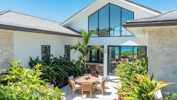 Uma casa moderna com uma grande janela de vidro, um pátio com mesa e cadeiras redondas e vegetação exuberante.