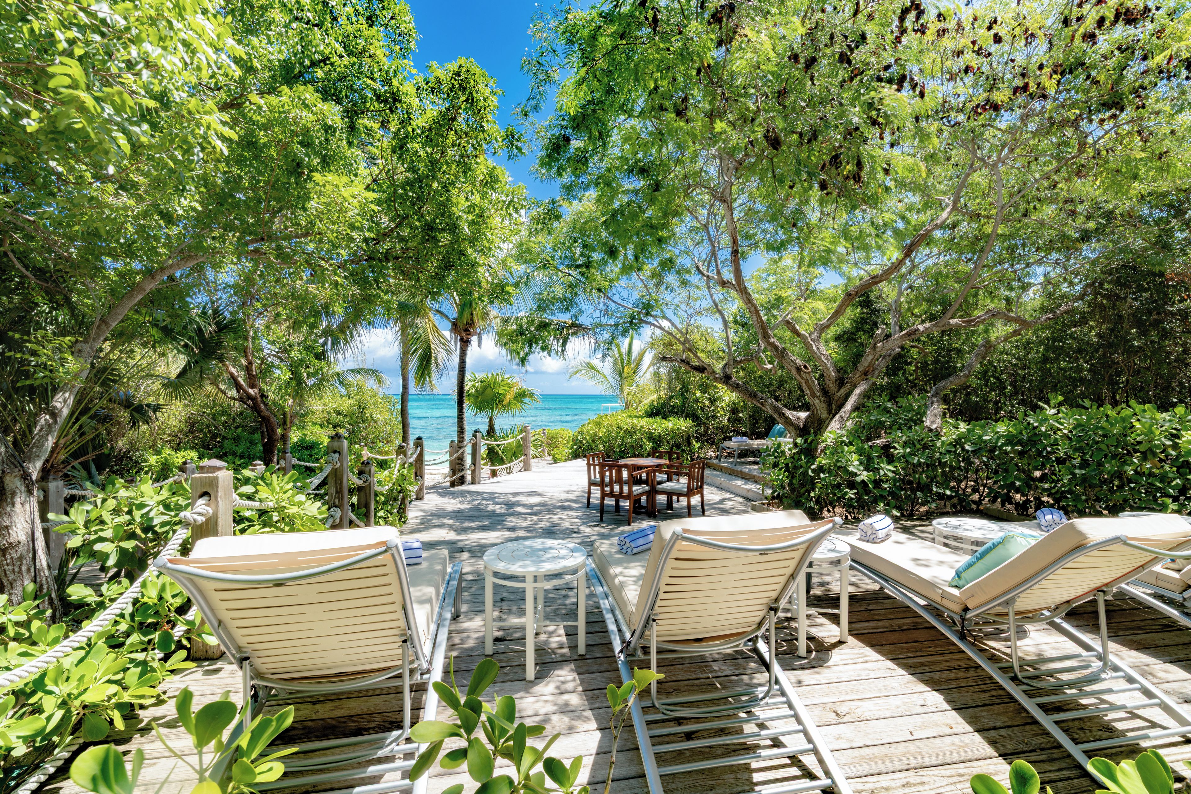 Une terrasse en bois, agrémentée de chaises longues et d’une table, entourée d’une végétation luxuriante et offrant une vue sur la plage.
