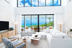 A modern living room with a large TV, comfortable seating, a dining area, and a view of the ocean.