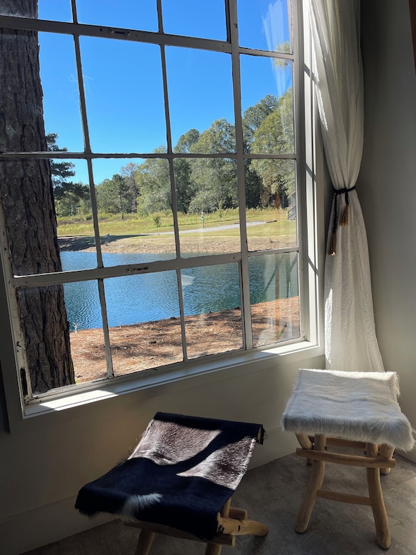 A room with a large window overlooking a body of water and a grassy area. There is a wooden stool with a fur throw and a white cushion.
