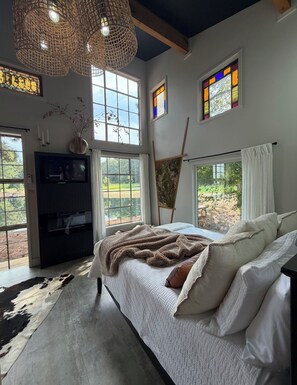 A bedroom with a large bed, a television, a painting, and a view of the outdoors.