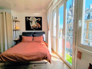 Un dormitorio con una cama, un cuadro de una vaca, una ventana con vistas a edificios y un cartel en la ventana.