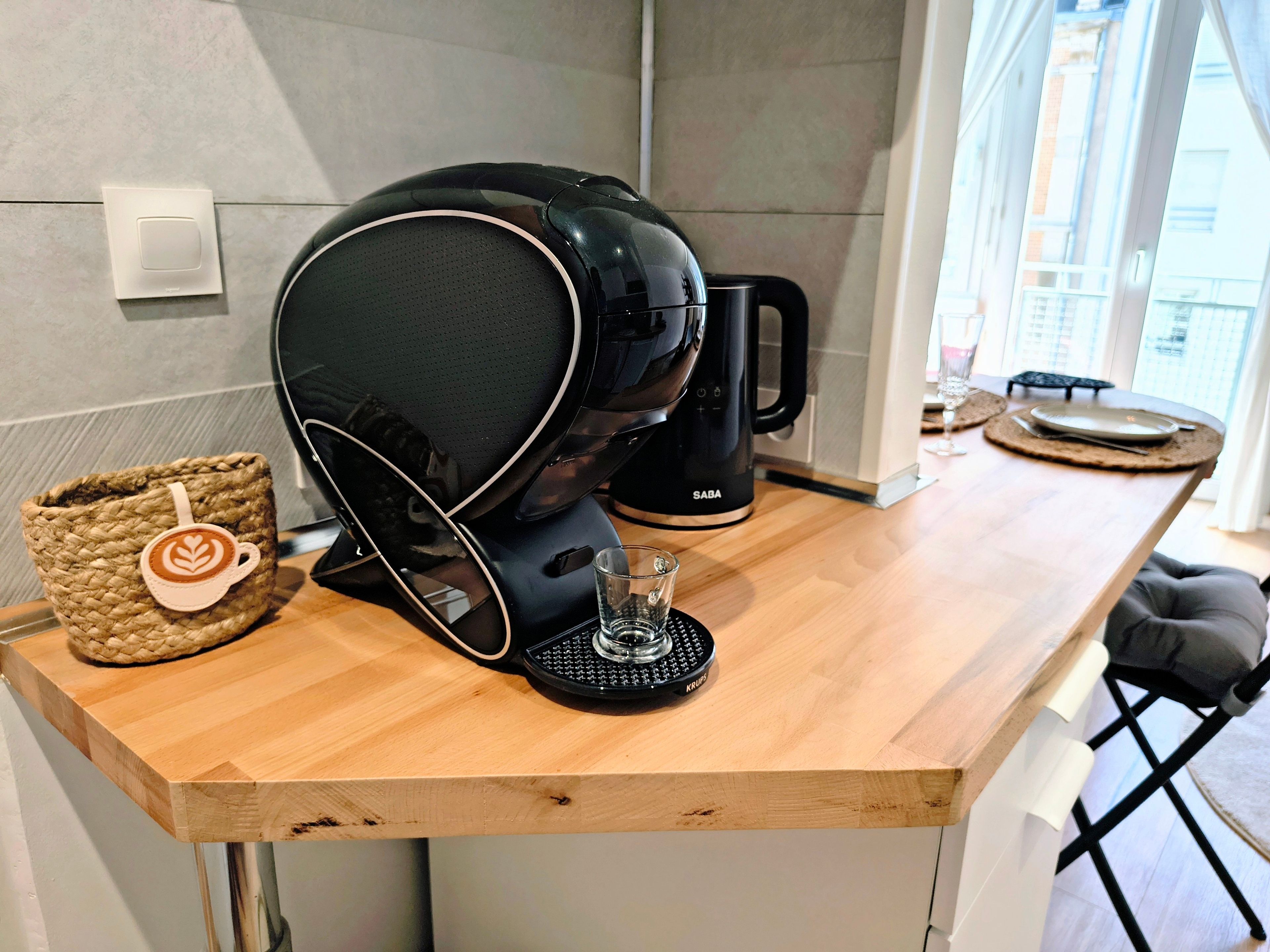 En kaffemaskin står på en bänkskiva av trä, bredvid en kaffekopp och ett glas.