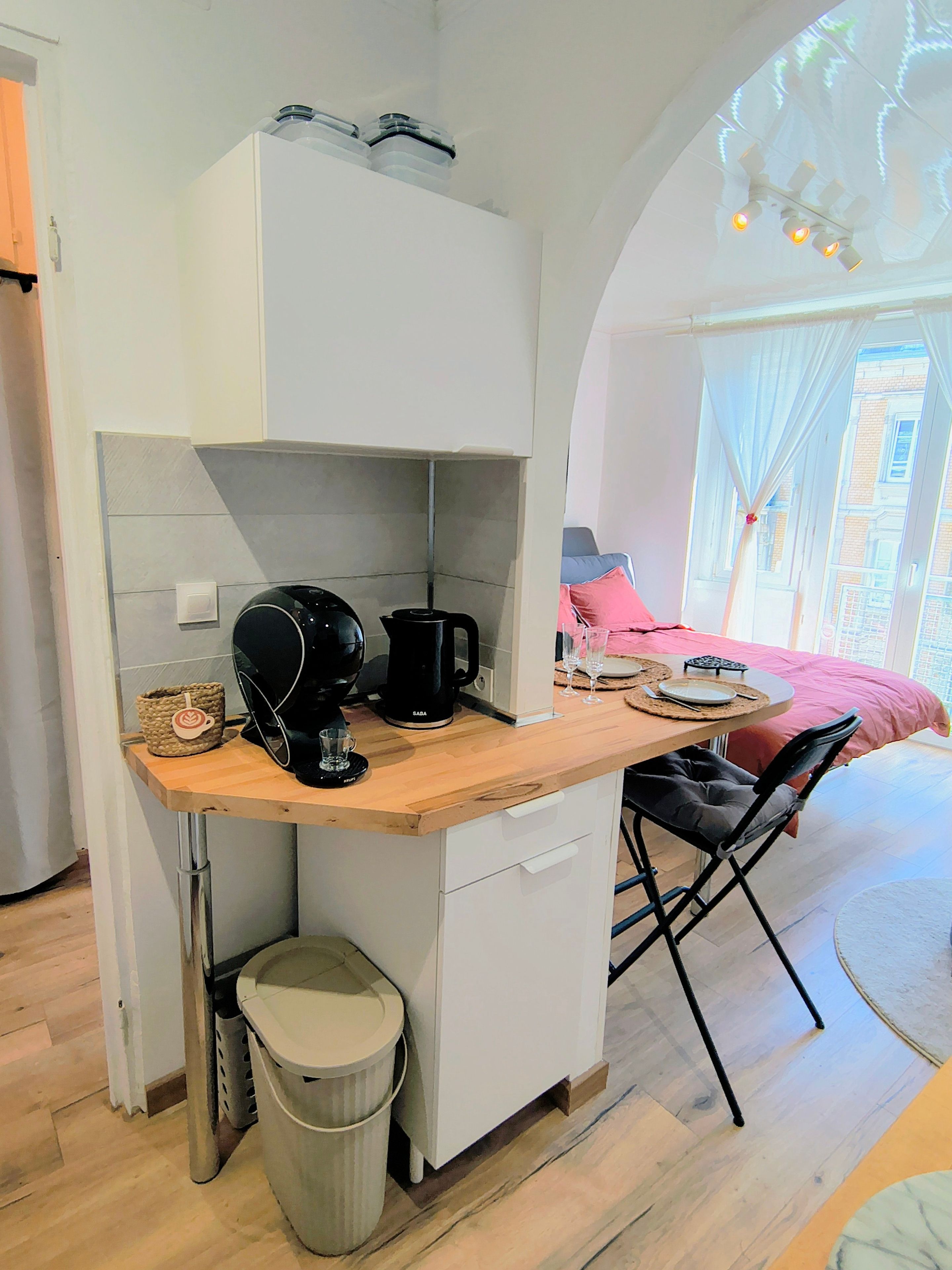 Eine kompakte Küche mit Kaffeemaschine, Mülleimer und kleinem Kühlschrank.
