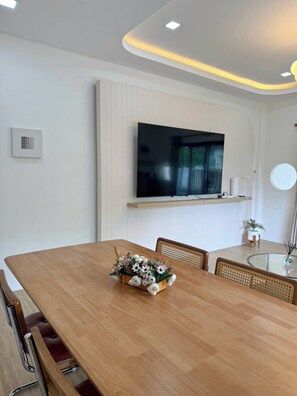 A modern dining area with a wooden table, chairs, a flat-screen TV mounted on the wall, and a small potted plant on the side. - 3BR near Old Town & Chillva Market - 8 Guests (Mueang Phuket)
