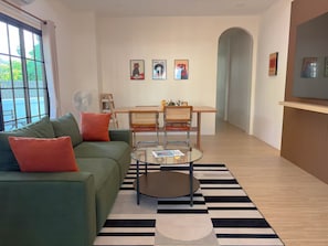 Un salon moderne avec un canapé vert, une table basse en verre et un tapis à rayures. On y trouve deux fauteuils et une petite table avec un ventilateur.