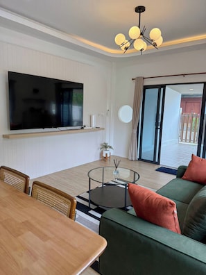 A modern living room with a flat-screen TV mounted on the wall, a round glass coffee table, a sofa with cushions, and a dining area with a wooden table and chairs. - 3BR near Old Town & Chillva Market - 8 Guests (Mueang Phuket)