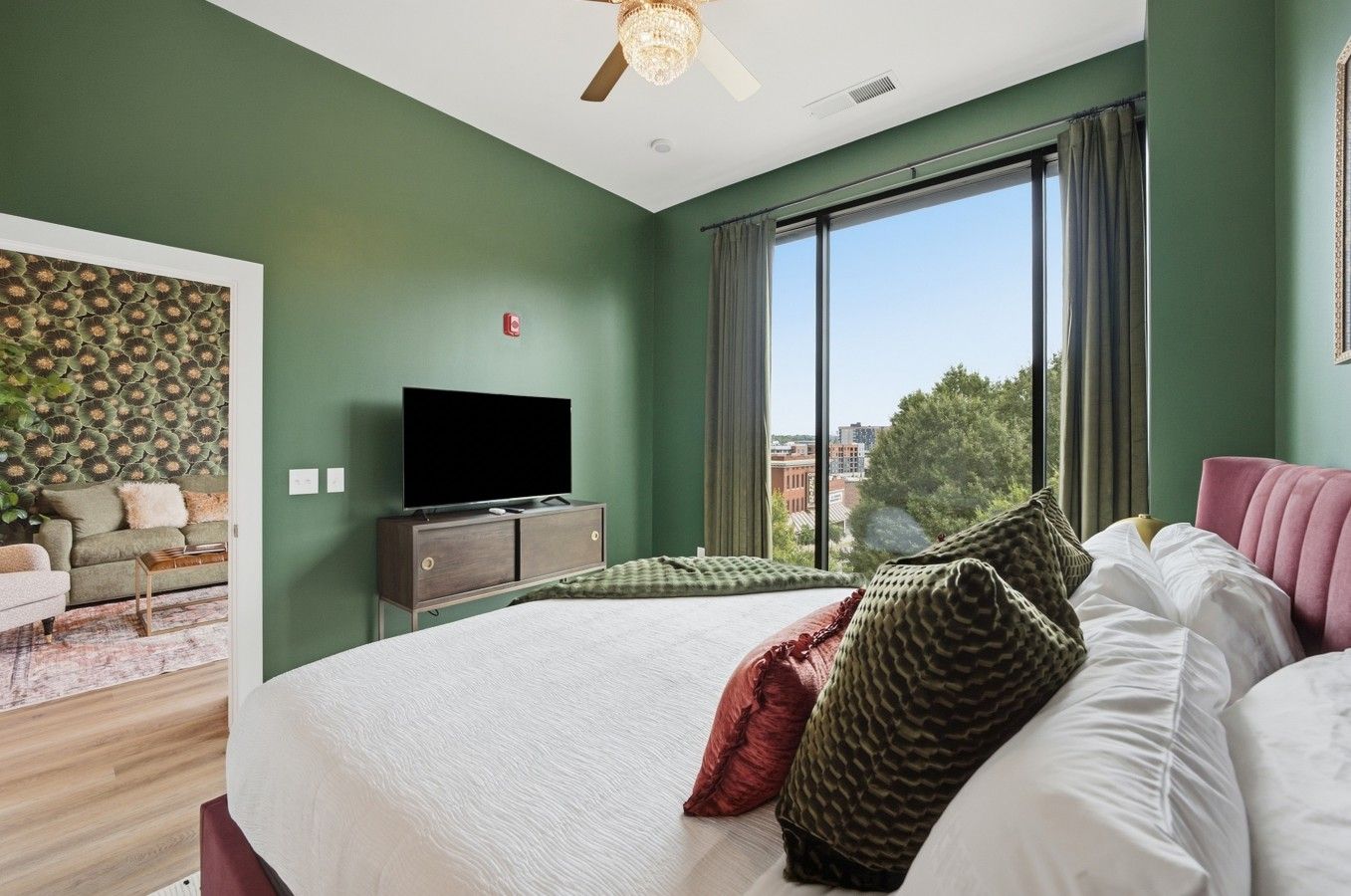 Une chambre à coucher avec un grand lit, un téléviseur posé sur un meuble, et une vue sur la ville grâce à de grandes fenêtres.