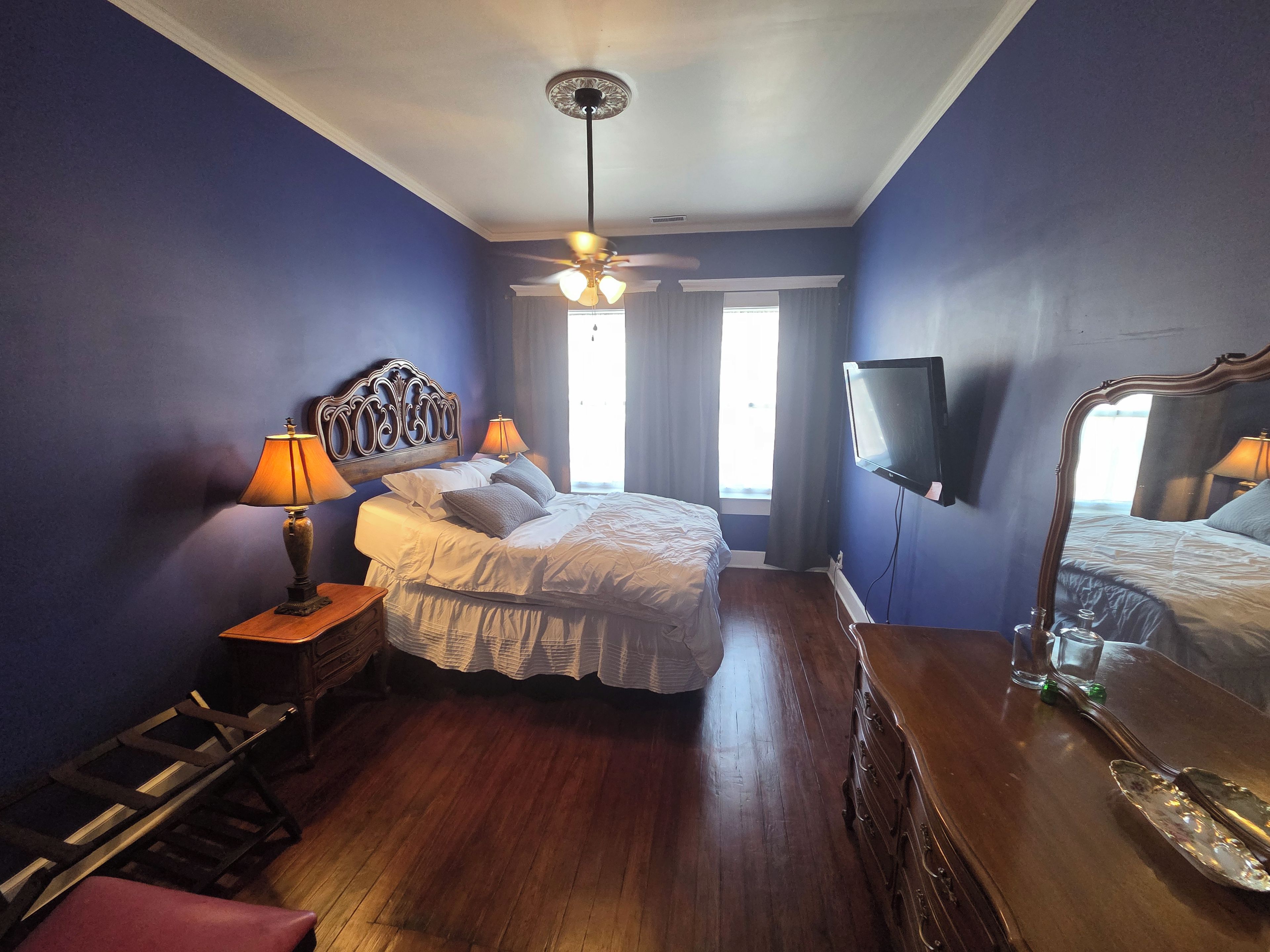 Une chambre à coucher comprenant un lit, deux lampes de chevet, un ventilateur de plafond, un téléviseur fixé au mur et une commode en bois.