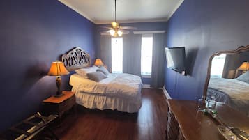 Une chambre à coucher comprenant un lit, deux lampes de chevet, un ventilateur de plafond, un téléviseur fixé au mur et une commode en bois.