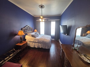 A bedroom with a bed, two bedside lamps, a ceiling fan, a TV mounted on the wall, and a wooden dresser.
