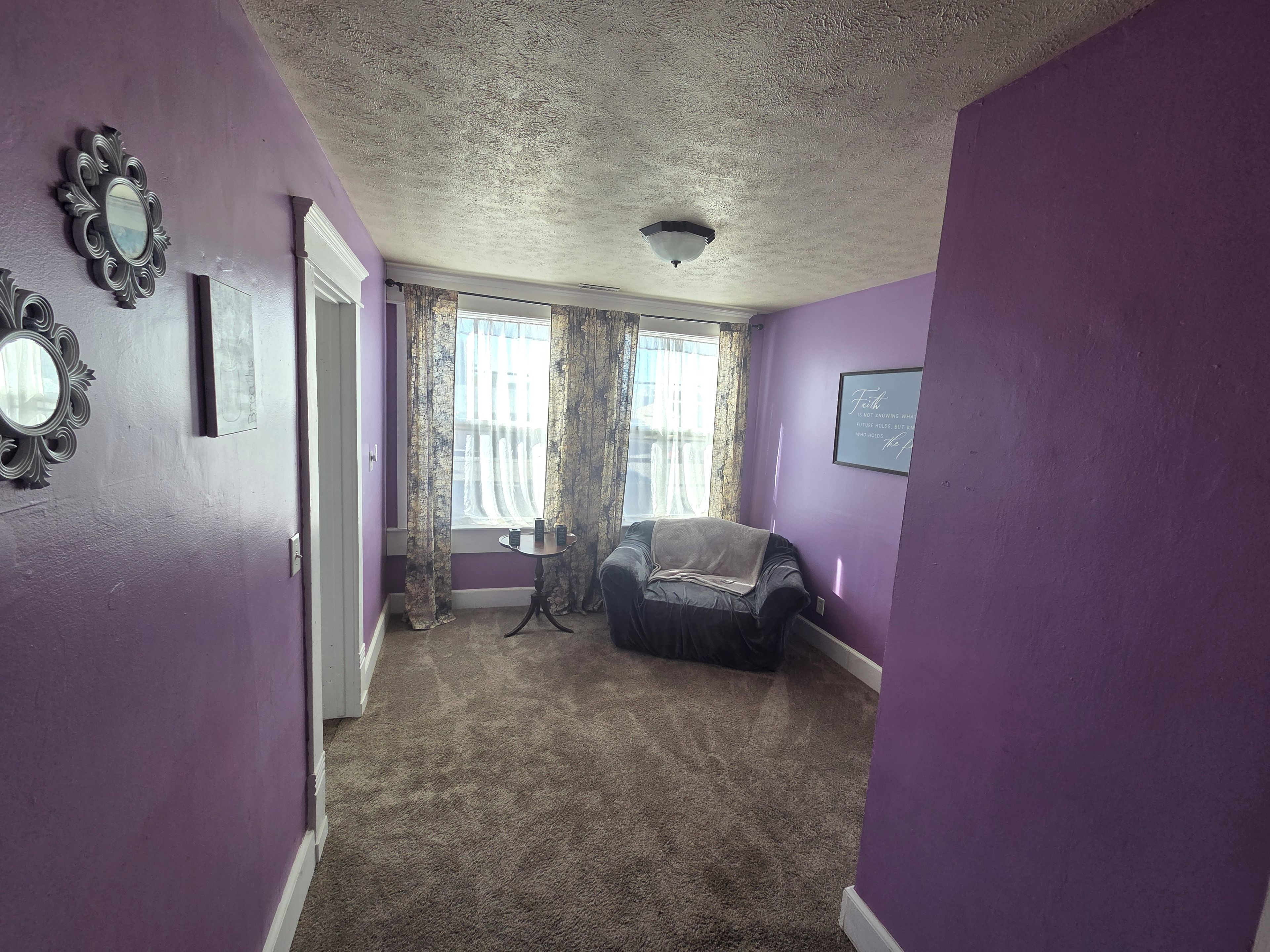 A room with purple walls, a ceiling light, a window with curtains, a small table, and a chair.