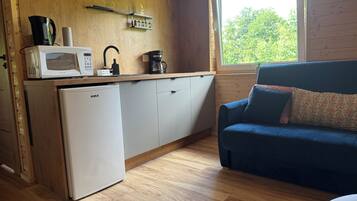 A compact kitchen with a microwave, coffee maker, and a small refrigerator. A blue sofa with patterned cushions is positioned near a window with a view of trees.