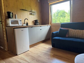 A compact kitchen with a microwave, coffee maker, and a small refrigerator. A blue sofa with patterned cushions is positioned near a window with a view of trees.