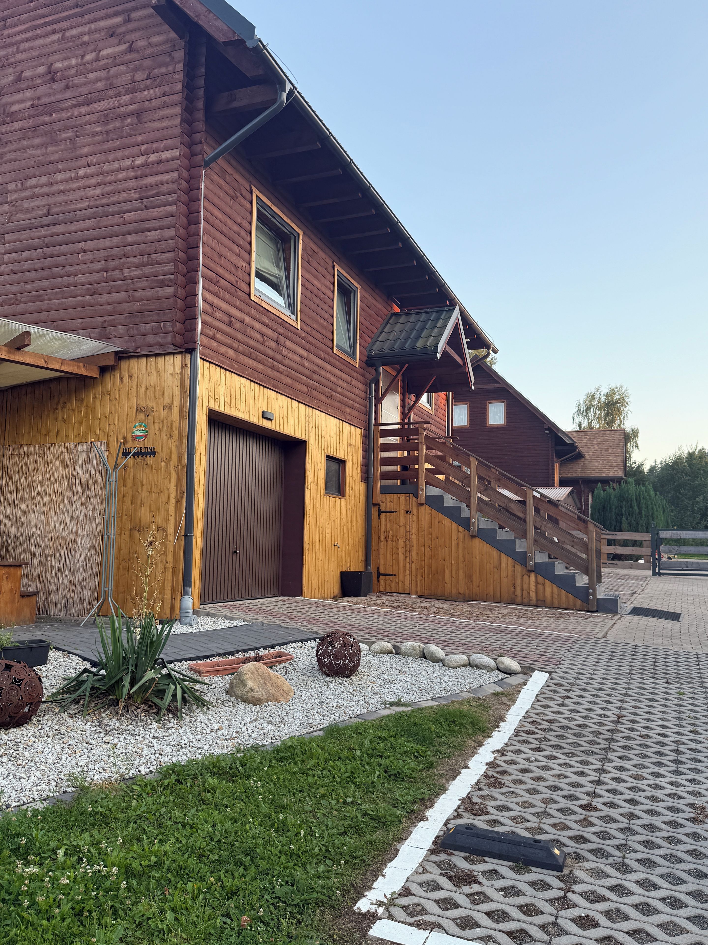 A wooden building with a garage, a staircase, and a paved area with plants and rocks.