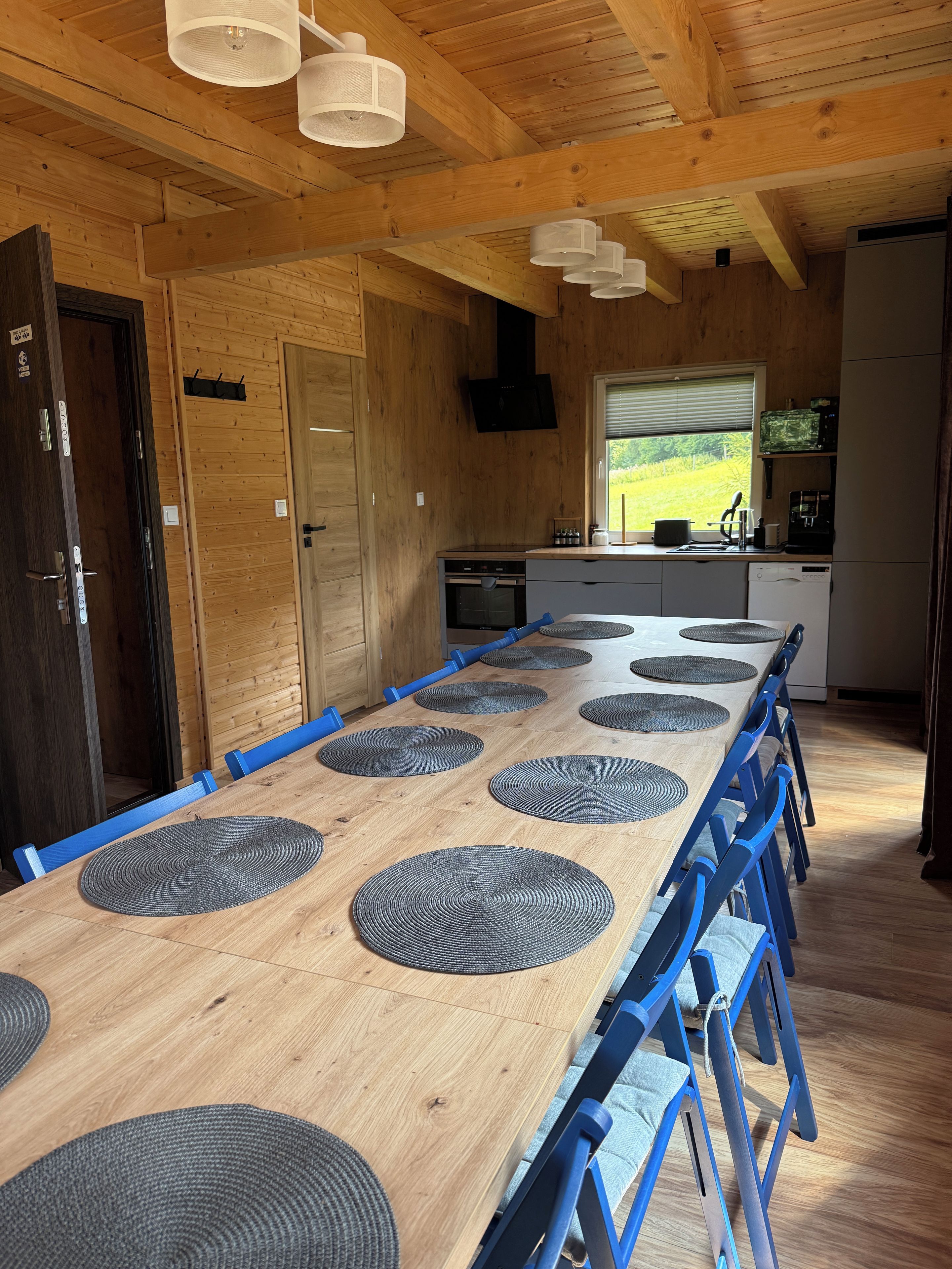 A wooden interior with a long dining table, blue chairs, and a kitchen area with appliances.