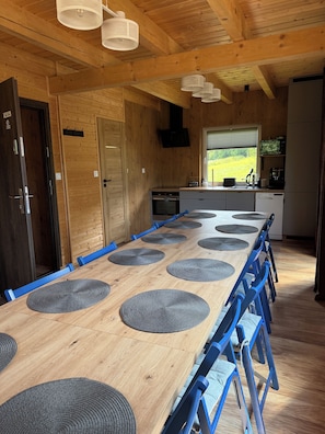 A wooden interior with a long dining table, blue chairs, and a kitchen area with appliances.
