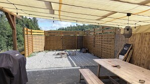 Un espacio al aire libre con patio, una mesa de madera y bancos, una parrilla y una pérgola con luces colgantes.
