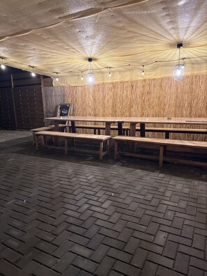 Ein Außenbereich mit Holzbänken und -tischen, einem Ziegelboden und einer Holzwand mit vertikalen Lamellen.