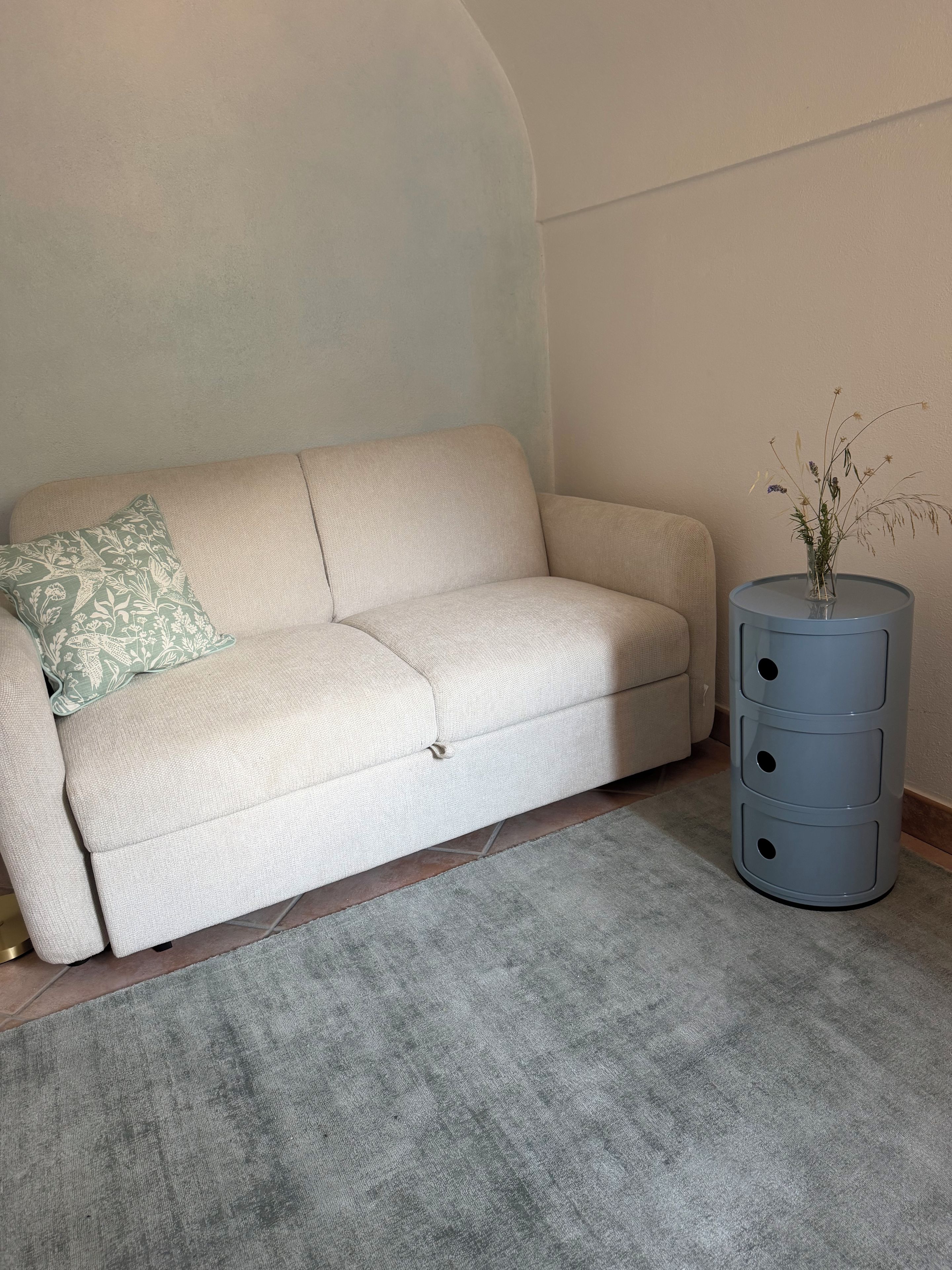 Un canapé beige agrémenté d’un coussin à motifs, un tapis gris et une petite table d’appoint cylindrique bleue sur laquelle est posé un vase de fleurs séchées.