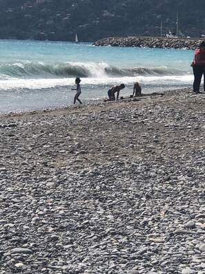 Une plage rocheuse avec des vagues, un voilier à l’horizon et des personnes près de l’eau.