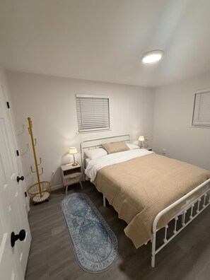 A bedroom with a bed, bedside tables, a lamp, a window with blinds, a coat rack, and a patterned rug. - Cozy 2 Bedroom Near Uptown, Dining & Fun (Charlotte)